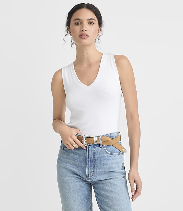 A woman in a white tank top and blue jeans with a tan belt stands against a plain white background.