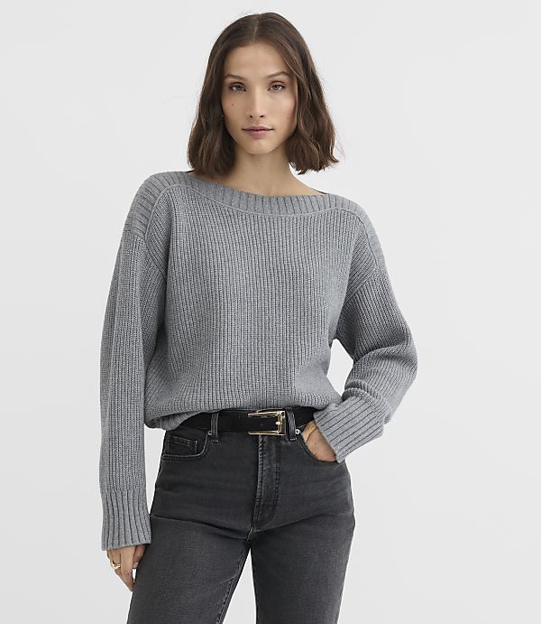 A woman in a grey ribbed sweater and dark jeans stands against a plain white background.