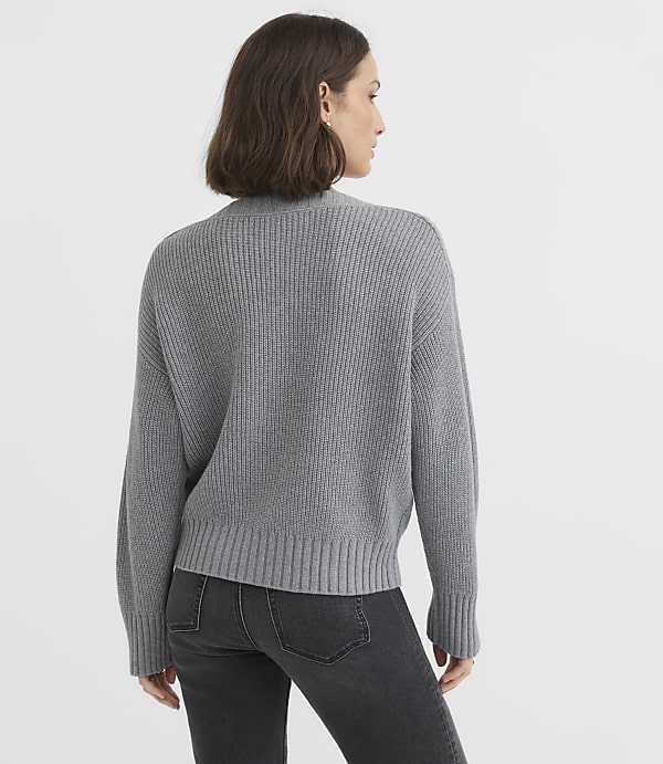 A woman in a grey sweater and jeans stands against a plain grey background.