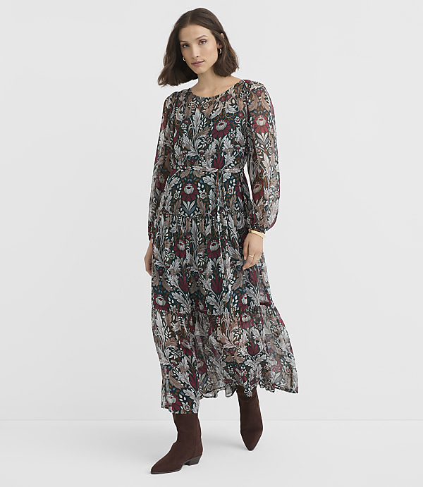 A woman in a long, patterned dress and brown boots stands against a plain white background.