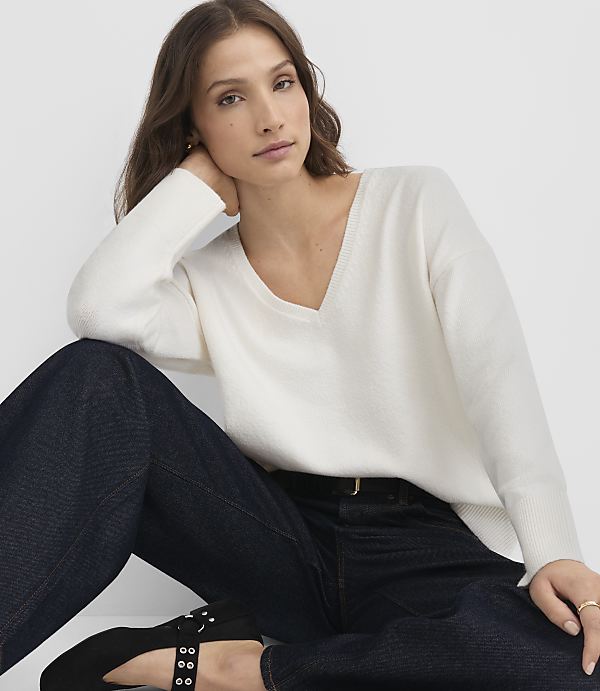 A woman in a white v-neck sweater and dark jeans poses against a plain white background.