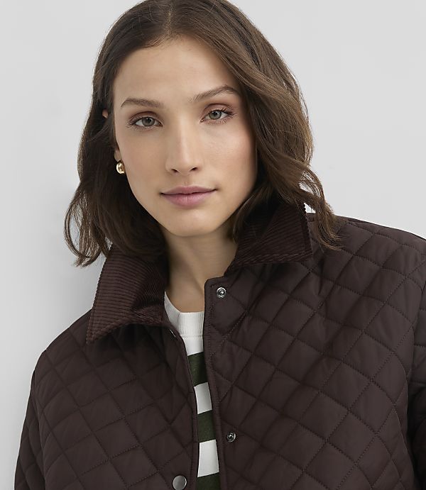 A woman with dark hair and light skin wears a brown quilted jacket with a corduroy collar, against a plain gray background.