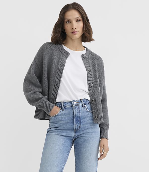 A woman in a grey cardigan and blue jeans stands against a plain grey background.