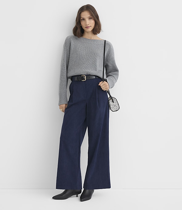 A woman in a gray sweater and navy pants stands against a plain white background.