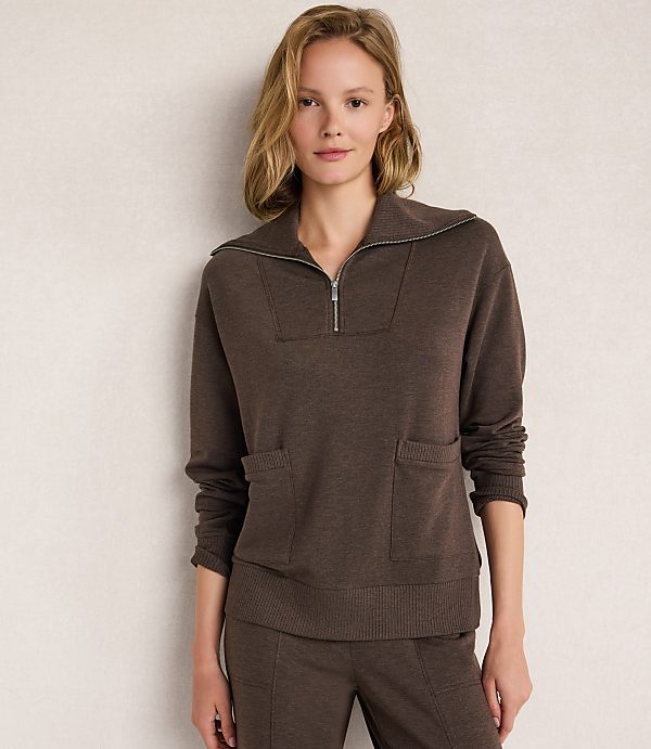 A woman in a brown quarter-zip sweater and matching pants stands against a plain, light-colored wall.