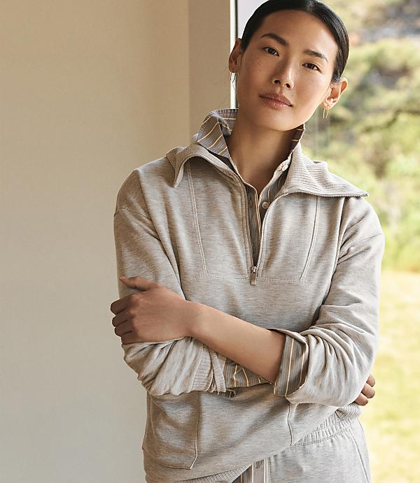A woman in a grey quarter-zip sweatshirt stands with her arms crossed in front of a softly blurred background of greenery and a window.