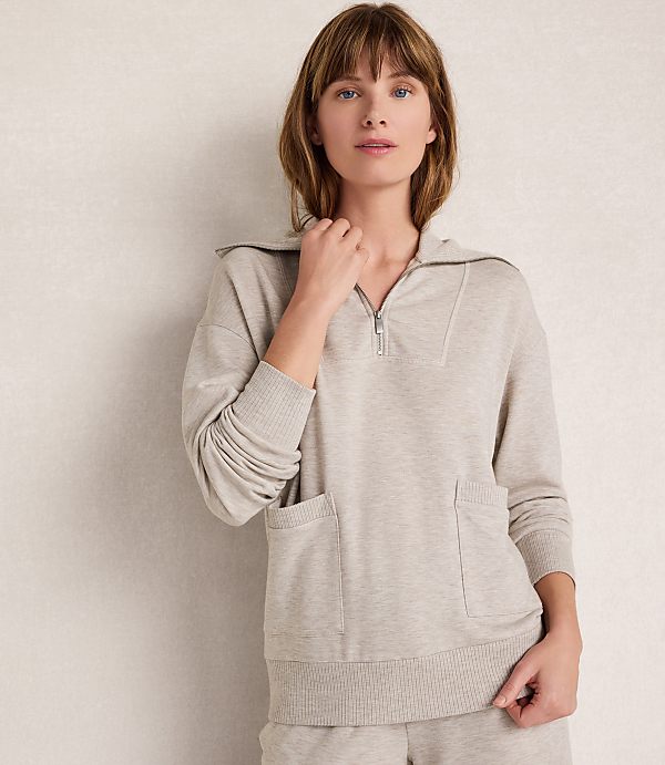 A woman in a light beige quarter-zip sweatshirt with pockets stands against a textured, light-colored wall.