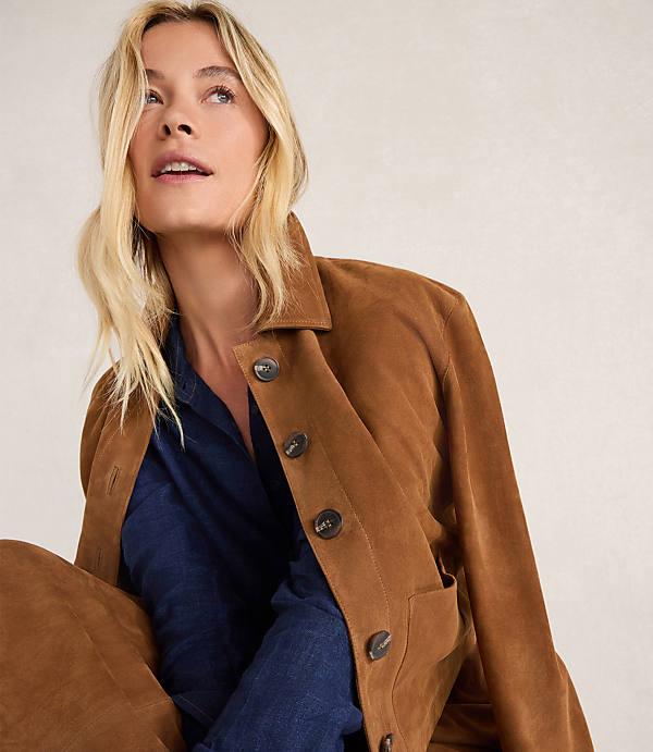 A blonde woman in a brown suede jacket and blue shirt looks up against a neutral background.