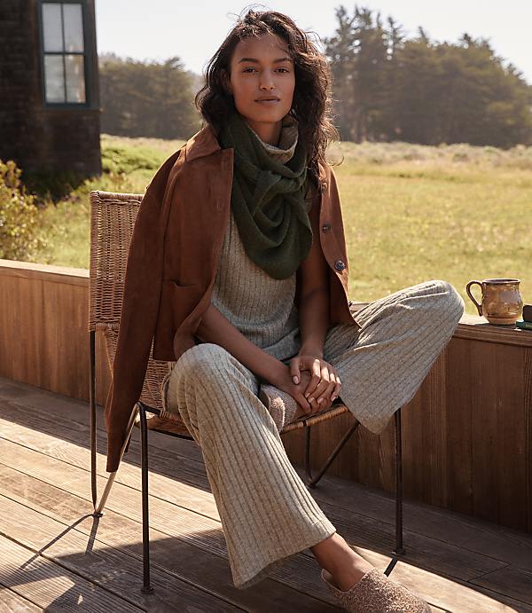 A woman in a brown jacket and green scarf sits on a wicker chair on a wooden deck, with a mug on the railing and a field of grass and trees in the background.