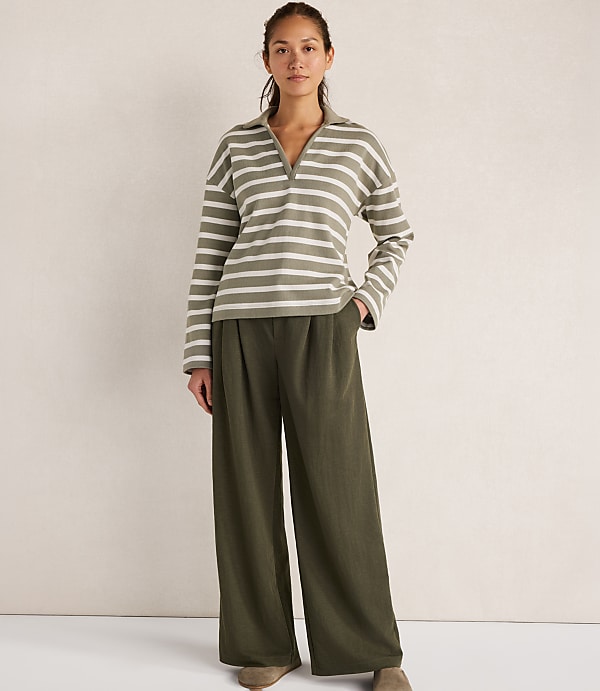 A woman in a striped collared shirt and wide-leg pants stands against a textured white background.