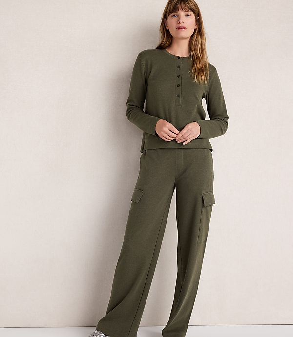 A woman in a green henley and cargo pants stands against a plain, light-colored wall.