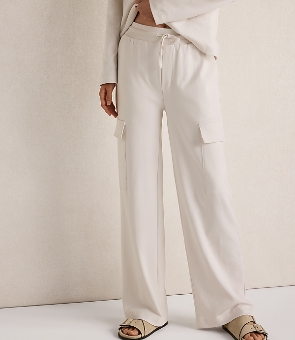 A person wearing cream-colored wide-leg cargo pants and sandals stands against a textured cream background.