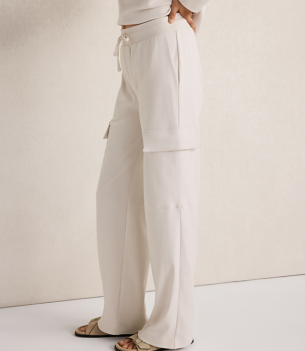 A person wearing cream-colored cargo pants and sandals stands against a textured cream wall.