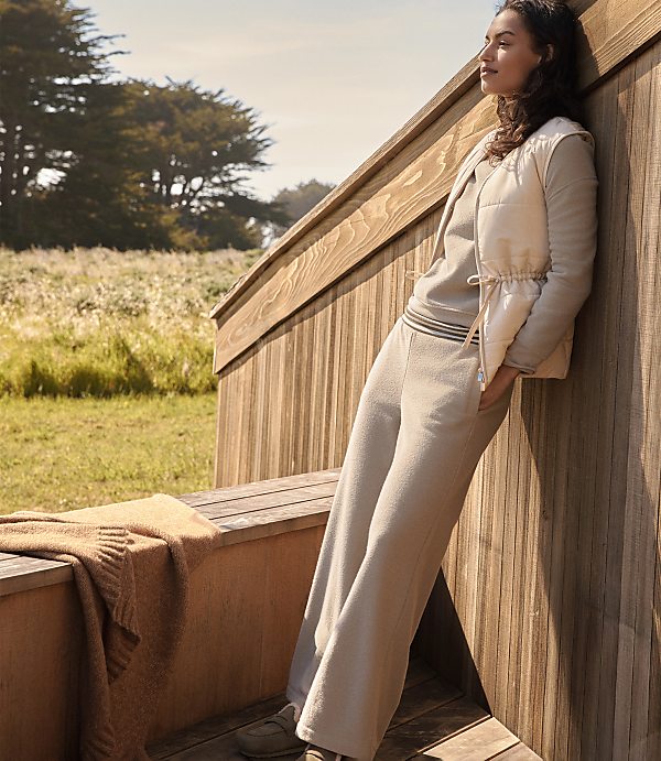 A woman in loungewear leans against a wooden structure with a sweater on a bench in the foreground and a grassy field with trees in the background.
