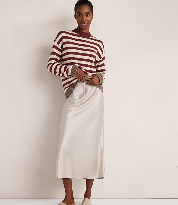 A woman in a striped sweater and satin skirt stands against a plain, light-colored wall.