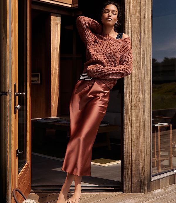 A woman in a rust-colored sweater and satin skirt leans against a wooden doorframe, with a blurred interior and outdoor scene in the background.