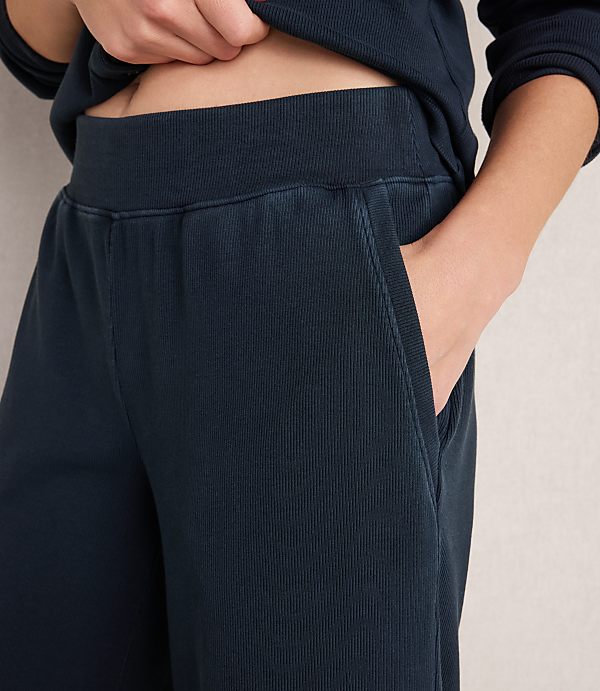 A person's hands are tucked into the pockets of dark blue ribbed sweatpants, with the waistband and a portion of a matching sweatshirt visible in the background.