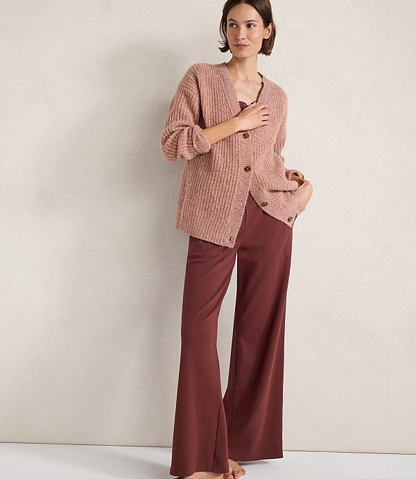 A woman in a pink cardigan and maroon pants stands against a plain, textured background.