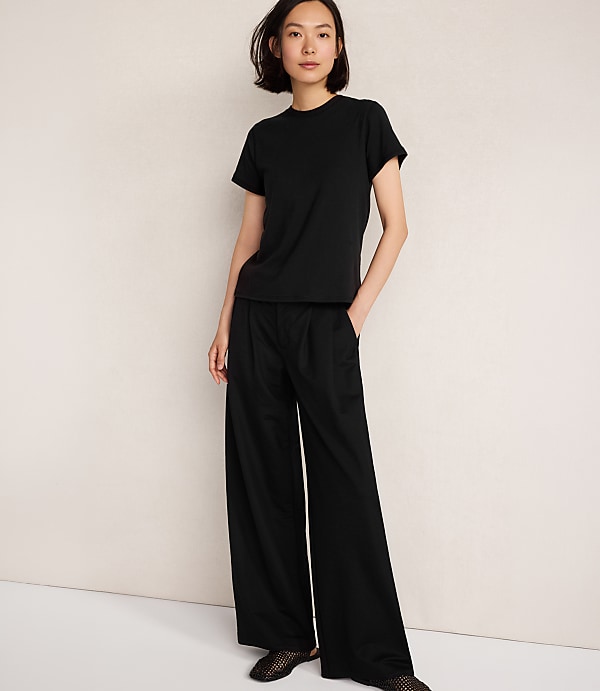 A woman in a black t-shirt and wide-leg pants stands against a plain, light-colored background.