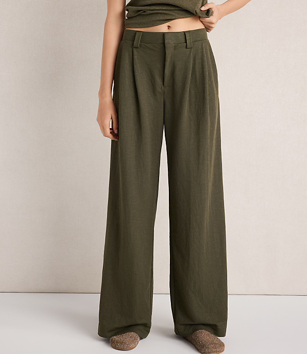 A person stands in olive green wide-leg pants and slippers against a textured, light-colored wall.