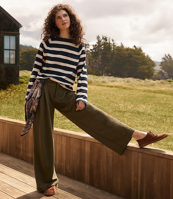 A woman in a striped shirt and olive pants poses on a wooden deck with a rustic cabin and a grassy field in the background.