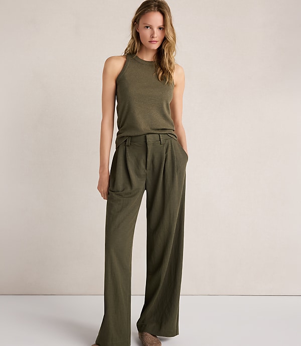 A woman in a green tank top and wide-leg pants stands against a plain, light-colored background.