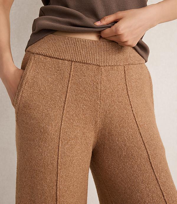 A person's hands are in the pockets of camel-colored knit pants, with a brown top visible above, against a plain background.