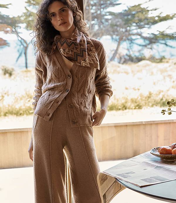 A woman in a brown cable-knit cardigan and matching pants stands in a sunlit room with a table in the foreground and a blurred outdoor scene in the background.