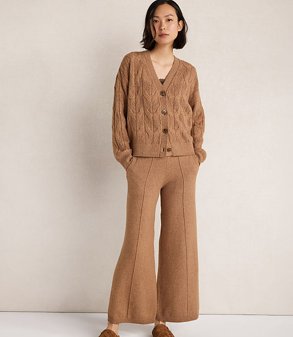 A woman in a brown cable-knit cardigan and wide-leg pants stands against a plain, light brown background.