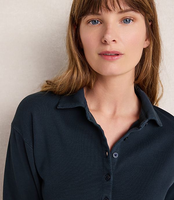 A woman with light skin and blue eyes, wearing a dark blue collared shirt, is shown against a plain, light-colored background.