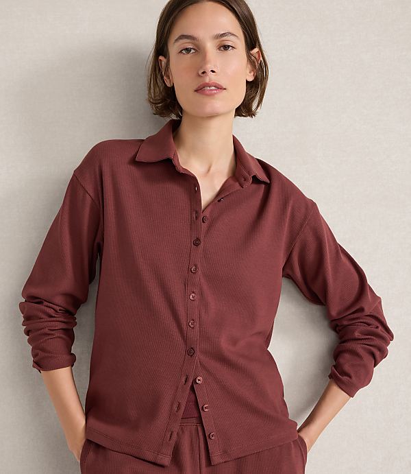 A woman in a maroon ribbed button-down shirt and matching pants stands against a textured gray wall.