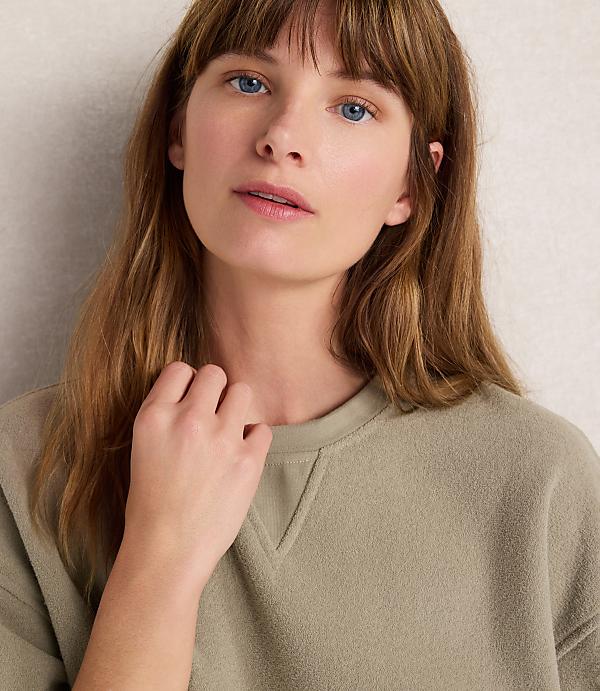 A young woman with light brown hair and blue eyes looks forward, her hand resting on her neck, against a plain, light-colored background.