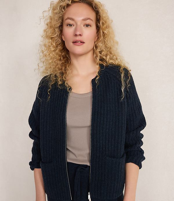 A woman with curly blonde hair wears a dark blue zippered cardigan over a taupe tank top, standing against a plain, light-colored background.
