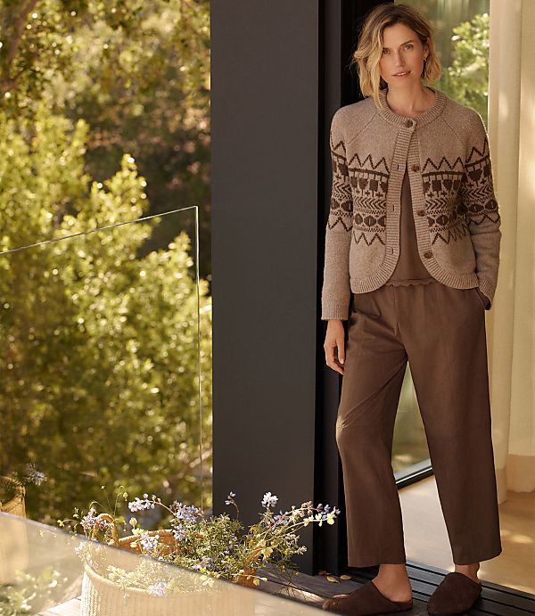 A woman in a patterned cardigan and brown pajamas stands by a window with lush green trees visible in the background and delicate flowers in the foreground.