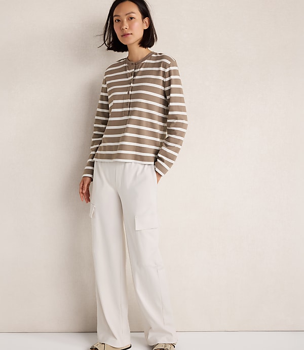 A woman in a striped long-sleeve shirt and white cargo pants stands against a textured cream wall.