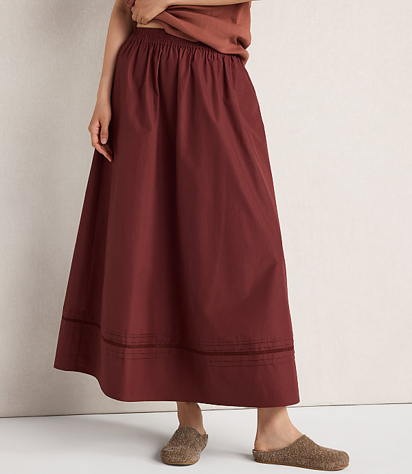 A person stands in a rust-colored skirt and brown clogs against a plain, light-colored background.