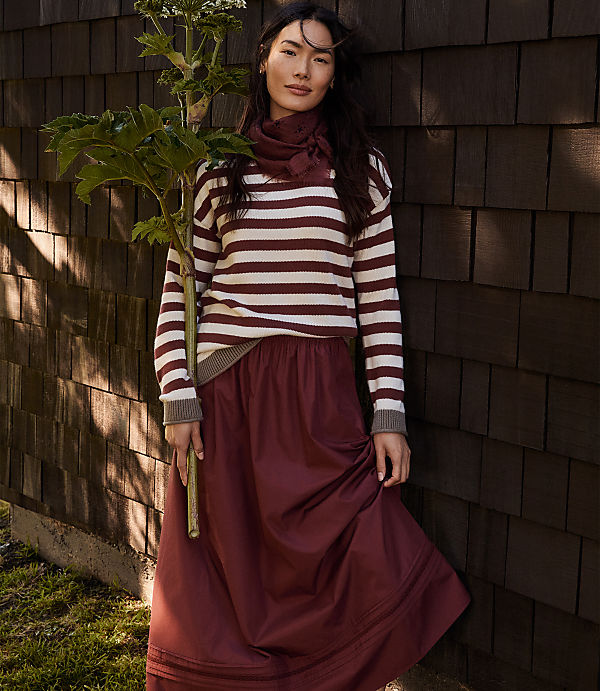 A woman in a striped sweater and maroon skirt stands in front of a wooden wall, holding a leafy green plant.