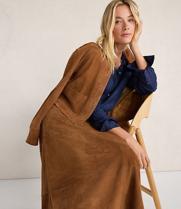 A blonde woman in a brown suede jacket and skirt sits on a wooden chair against a plain background.
