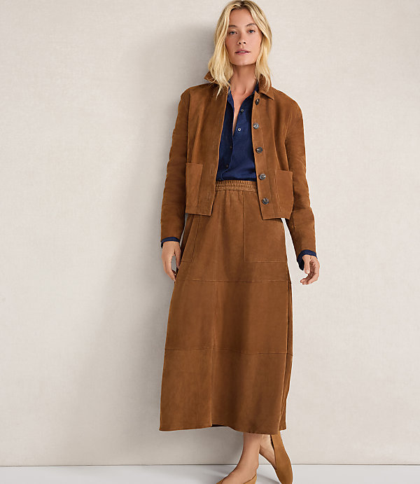 A woman in a brown suede jacket and skirt stands against a plain, light-colored wall.