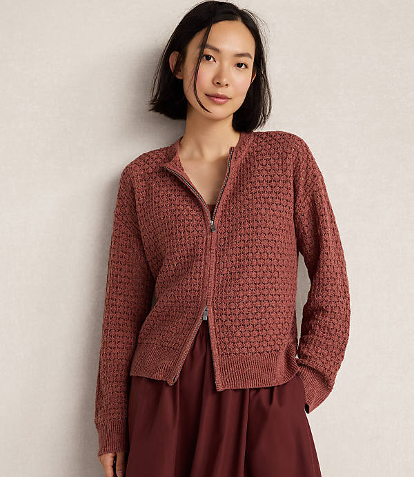 A young woman in a textured rose-colored zip-up cardigan stands against a plain, light-colored wall.