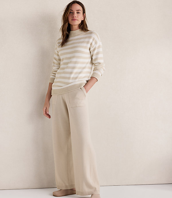 A woman in a striped sweater and wide-leg pants stands against a plain, textured wall.