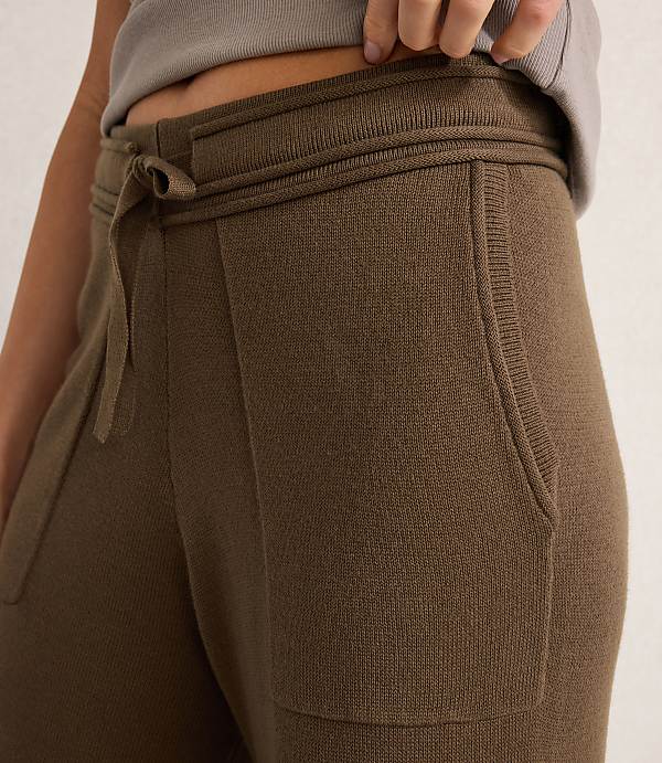 A close-up of olive green knit pants with a drawstring waist and pockets is shown against a blurred, neutral background.