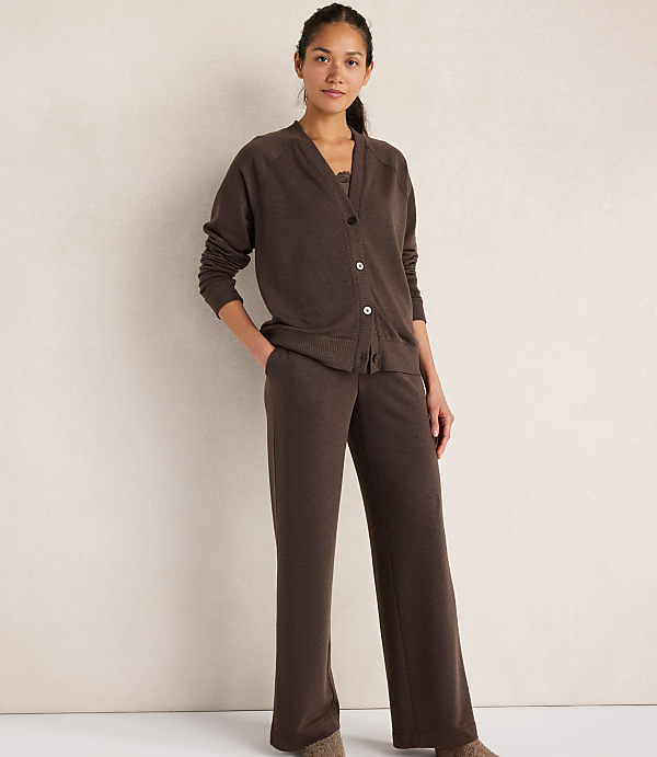 A woman in a brown cardigan and matching pants stands against a plain, light-colored wall.