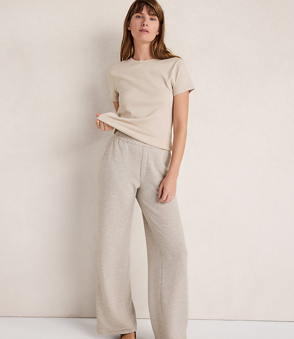 A woman in a beige t-shirt and light gray sweatpants stands against a textured, off-white background.