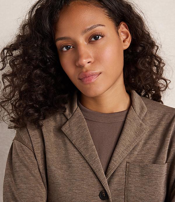 A woman with curly dark hair wears a brown jacket over a ribbed brown shirt, with a plain, light-colored background.
