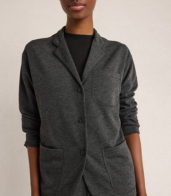 A woman in a dark grey blazer stands against a plain, light grey background.