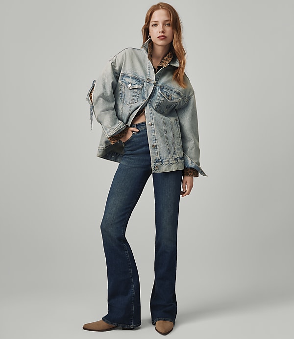 A woman in a denim jacket and flared jeans stands against a plain gray background.