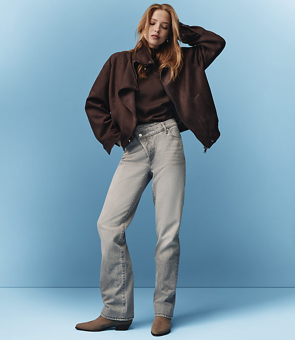 A woman in a brown jacket and light gray jeans stands against a solid blue background.