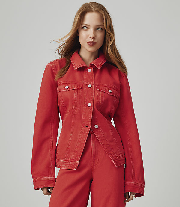 A young woman with red hair and a red denim jacket and pants stands against a plain gray background.