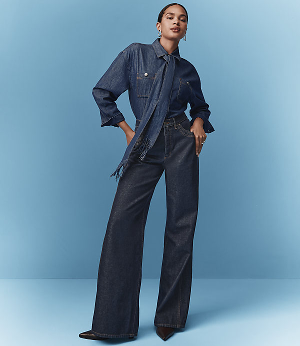 A woman in a denim shirt and wide-leg jeans poses against a solid blue background.